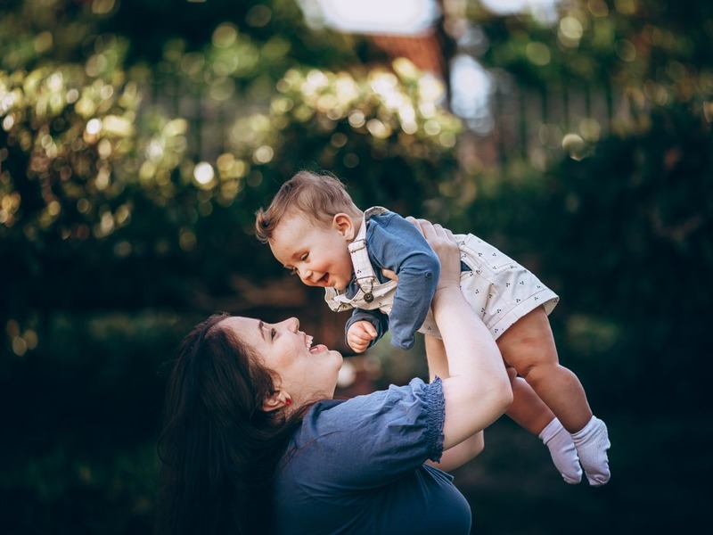 Mother joyfully lifting her laughing baby outdoors in the sunshine - Harmony Surrogacy